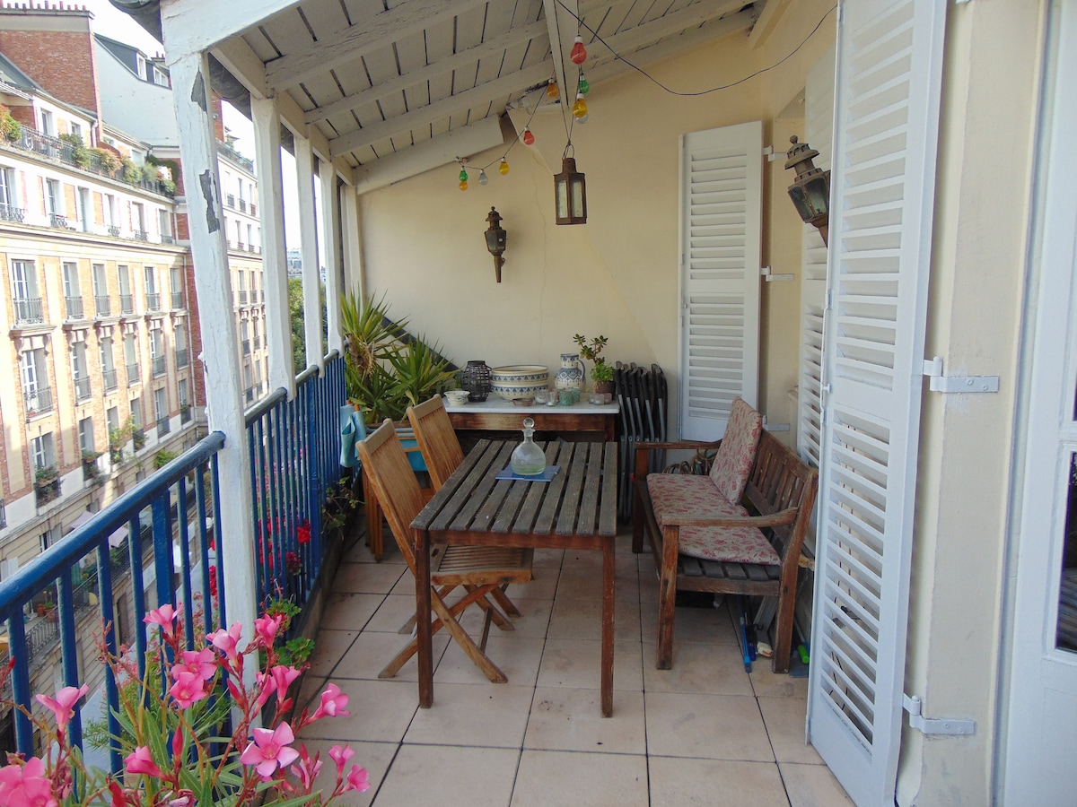 A charming terrace on the last floor features a wooden dining table surrounded by folding chairs and a bench. Potted plants add greenery, and colorful lights are strung above. The terrace offers an open view of the surrounding Paris rooftops.