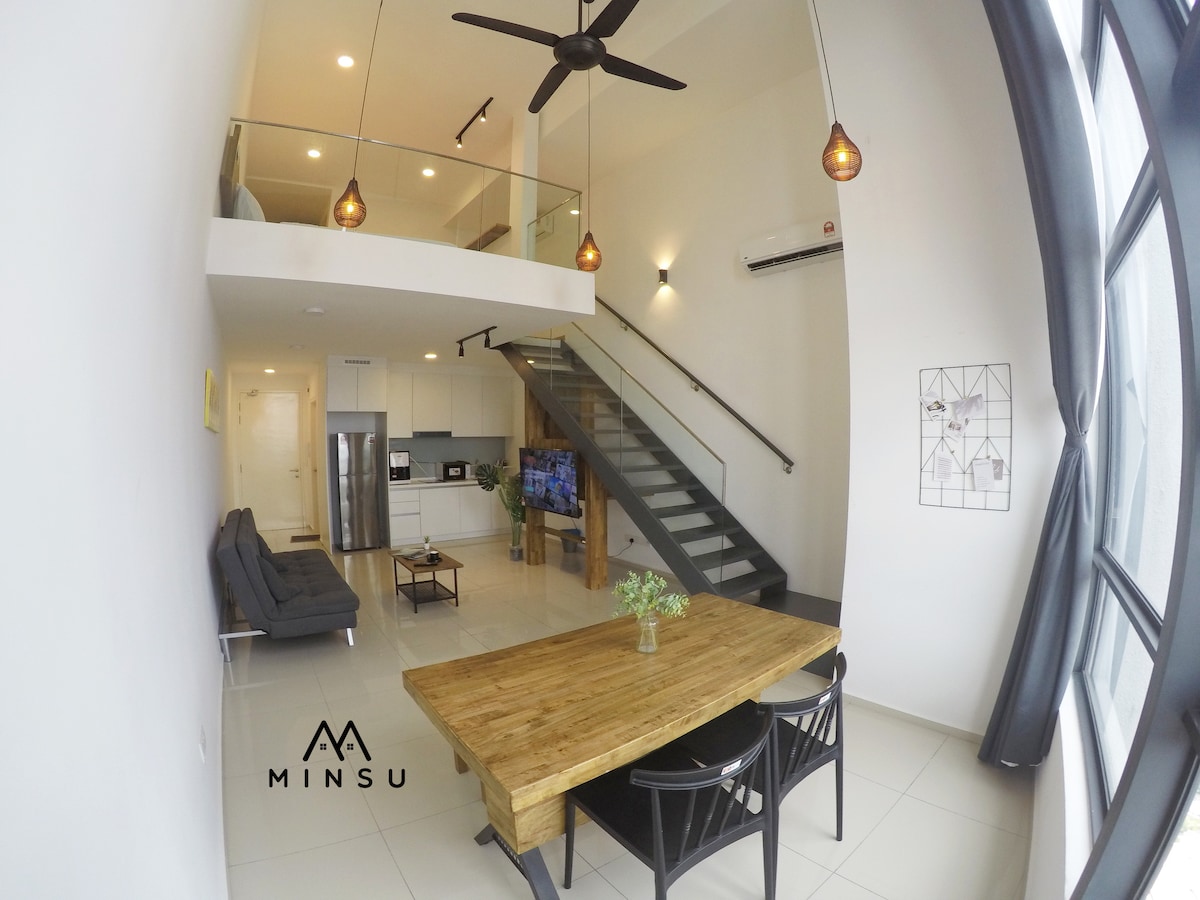 An open-plan living area is highlighted by a wooden dining table surrounded by black chairs. A sleek staircase leads to the upper level. Natural light streams through large windows, and a cozy seating arrangement is present, providing a modern and spacious ambiance.