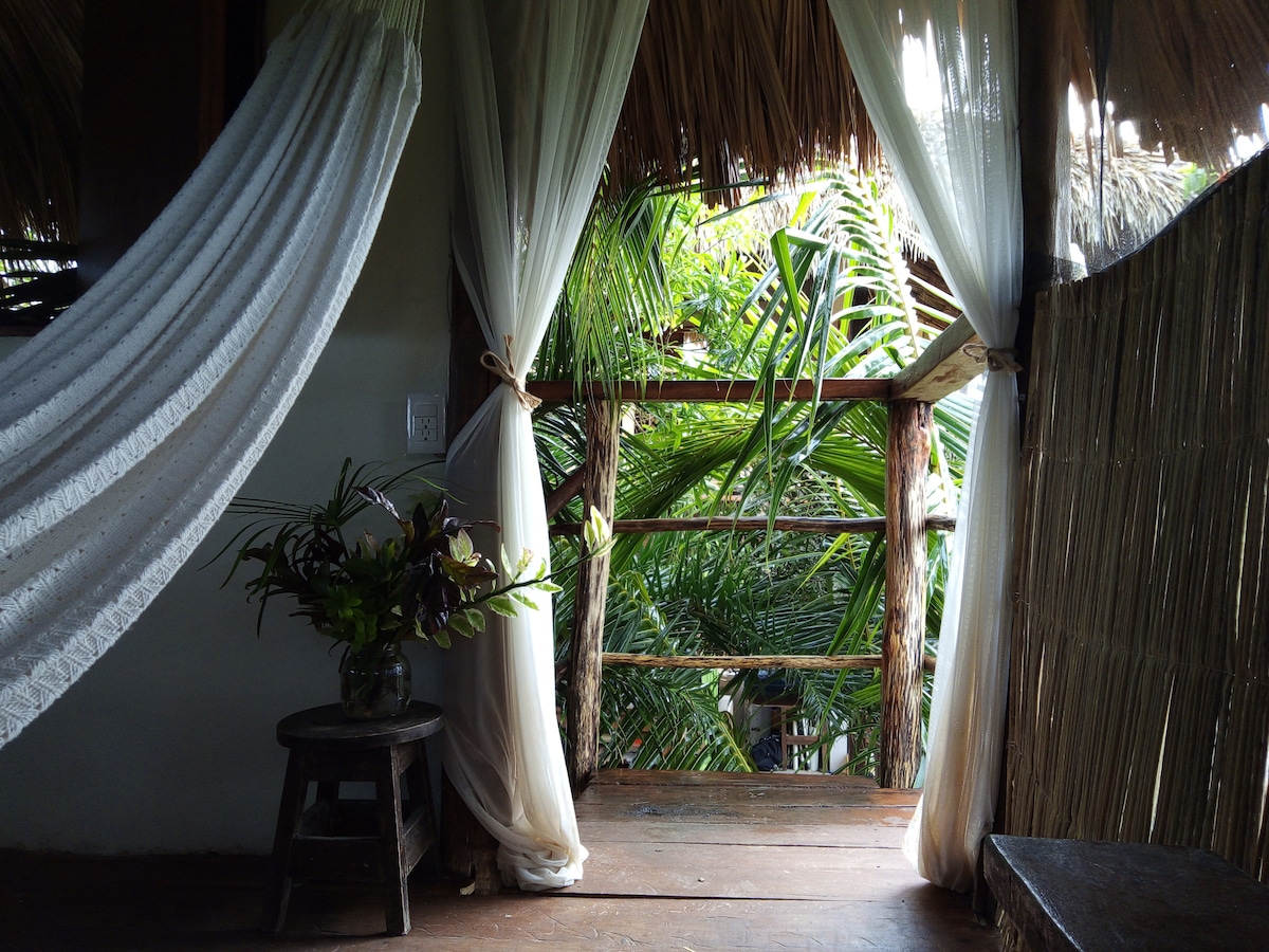 A doorway framed by flowing white drapes opens to a lush view of the surrounding greenery. A small wooden stool sits nearby on the polished floor, complementing the natural ambiance created by the palm trees visible through the entrance.