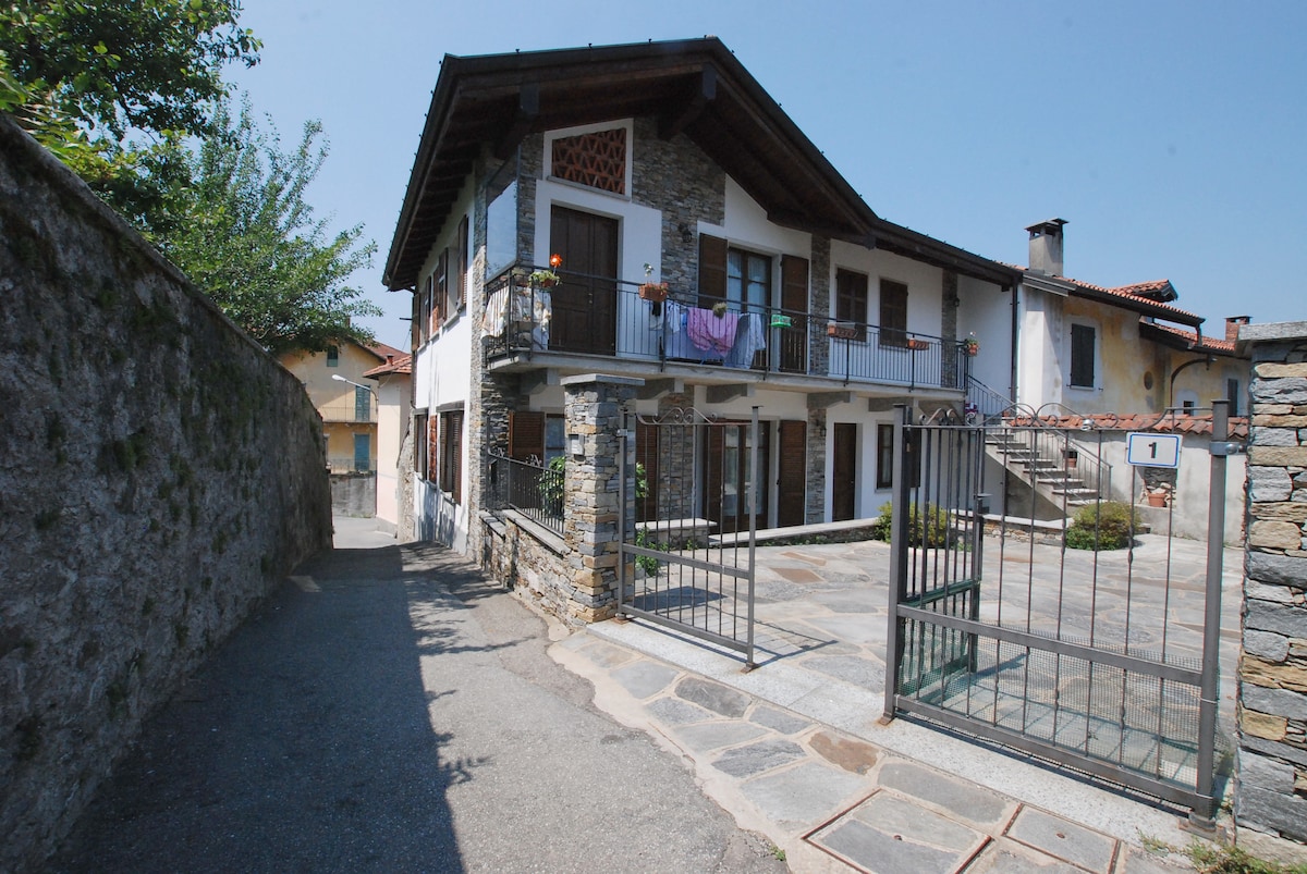 A characterful stone house is depicted, featuring a two-story structure with wooden balconies. The entrance is enclosed by a metal gate, and a cobblestone path leads to the courtyard. Colorful laundry hangs drying on the balcony, surrounded by greenery.