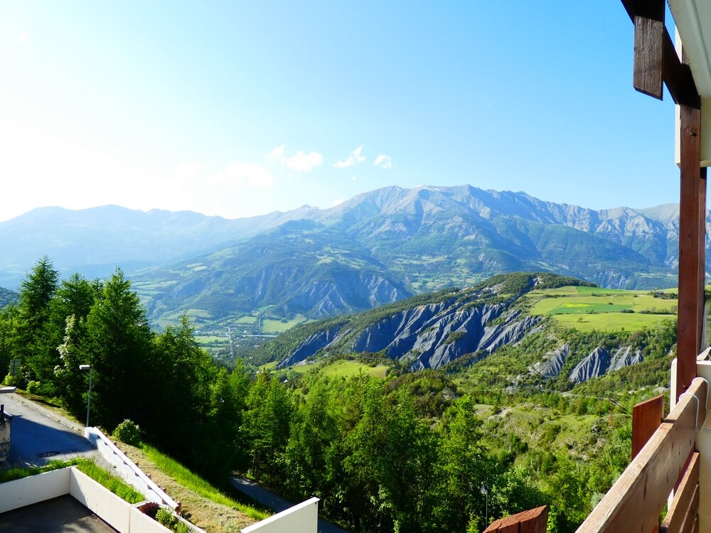 A panoramic view showcases rolling green hills and rugged mountain ranges under a clear blue sky. Natural landscapes extend into the distance, with an intriguing mix of steep slopes and valleys, creating a serene backdrop for a mountain getaway.