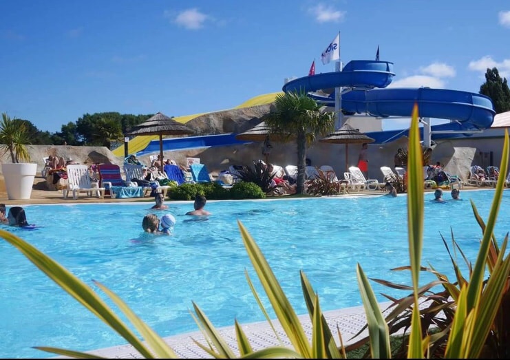 A vibrant swimming pool is surrounded by lounge chairs and tall palm plants. Water slides in blue and yellow are visible in the background. Guests are enjoying the water, while umbrellas offer shade in the sunny environment.