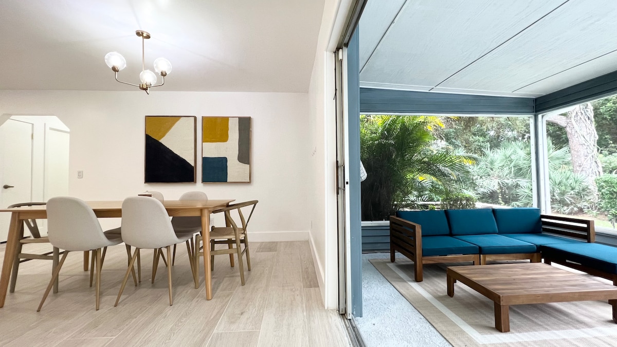 A dining area features a wooden table surrounded by four light-colored chairs. A pair of abstract wall art pieces adds color to the space. Adjacent to it, a screened porch showcases a teal sectional sofa and a wooden coffee table, with lush greenery visible outside.