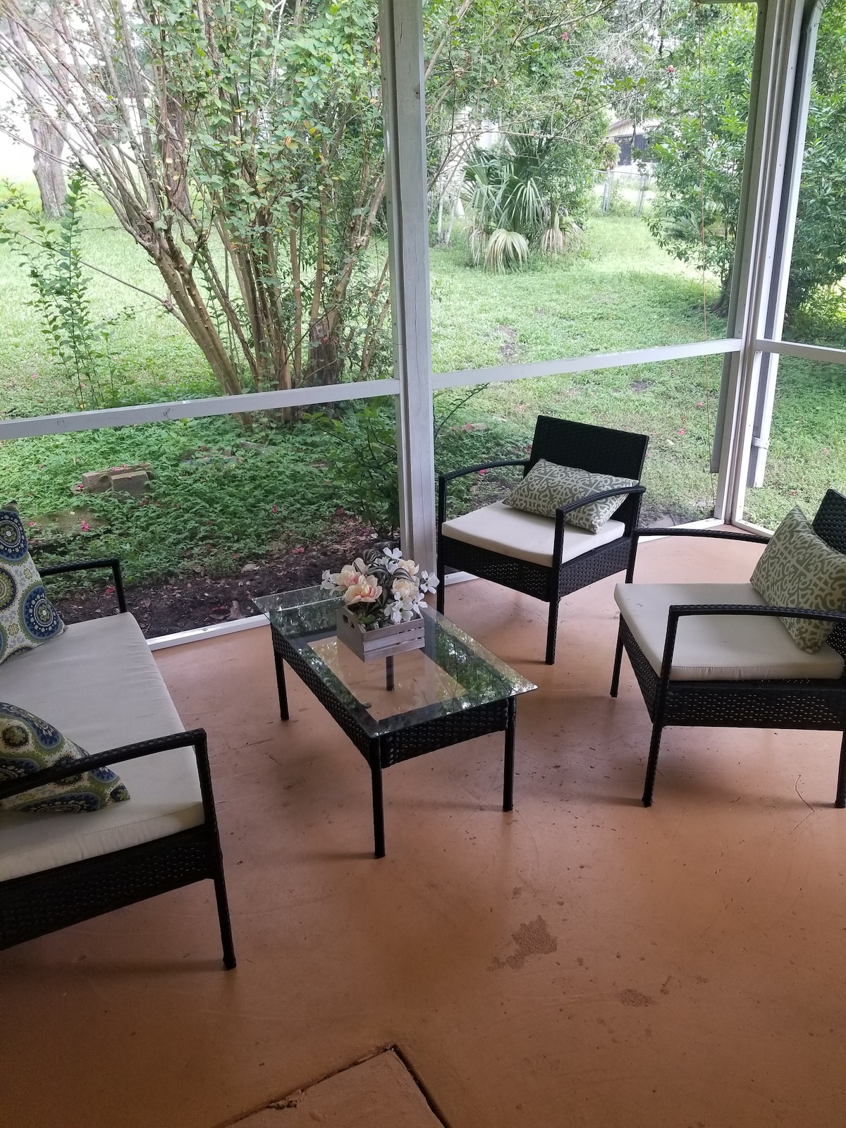 A screened Florida room contains four cushioned chairs arranged around a glass-top table. Decorative pillows add color while a small vase of flowers rests on the table. Lush greenery is visible through the surrounding screens, enhancing the inviting space.