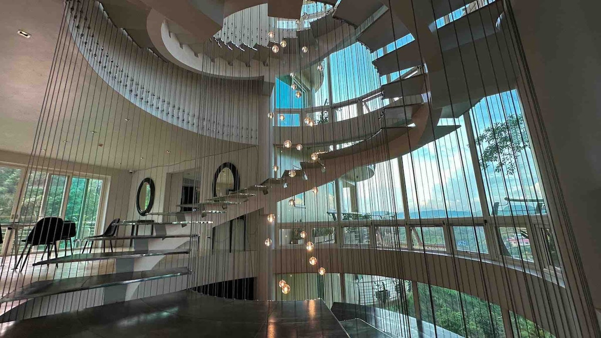 A modern spiral staircase is surrounded by glass and soft lighting, creating an open and airy feel. The staircase features flat steps and elegant railings. Large windows allow natural light to fill the space, offering views of the surrounding greenery.