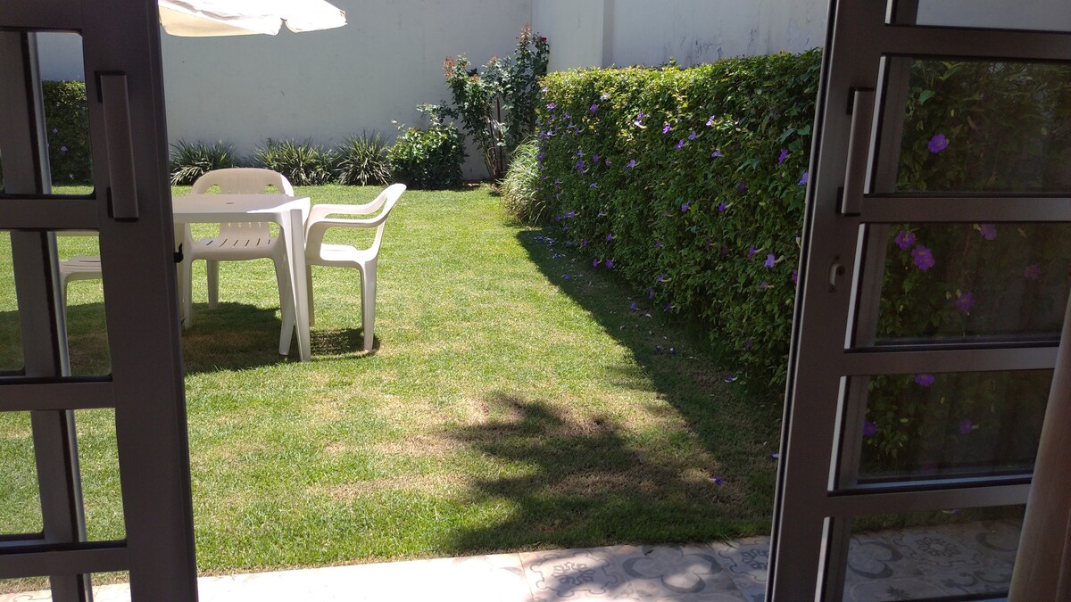 A well-maintained garden area is visible, showcasing a lush green lawn bordered by hedges and flowering plants. A white table and chairs are arranged on the grass, providing an inviting space for outdoor relaxation.