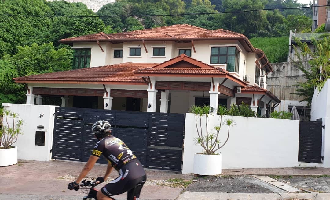 A spacious two-story house with a red-tiled roof is nestled among green hills. White walls are accented by a black gate and planters. A cyclist passes by, enjoying the peaceful surroundings. Lush greenery can be seen in the background, enhancing the serene atmosphere.