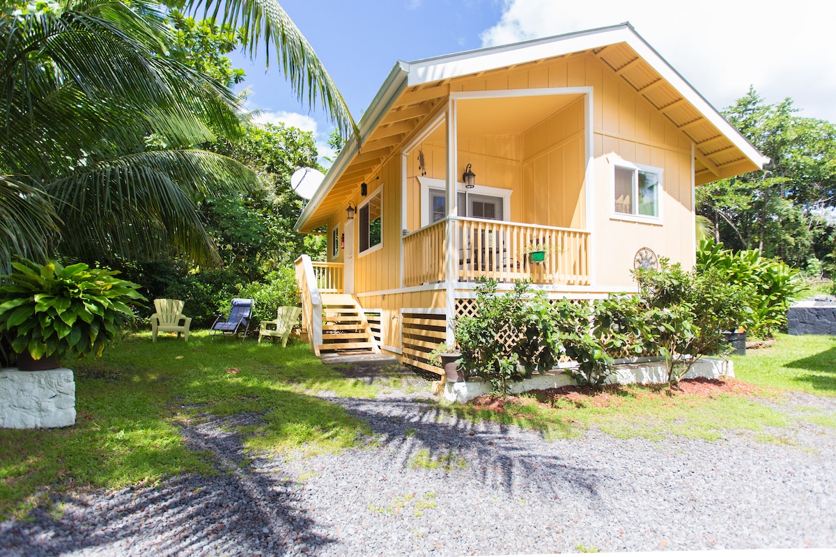 A charming yellow cottage is set amidst lush greenery, accessible via a wooden staircase. Adirondack chairs are positioned on the lawn, inviting relaxation. The home features a spacious front porch with a welcoming entry, complemented by tropical landscaping surrounding the property.