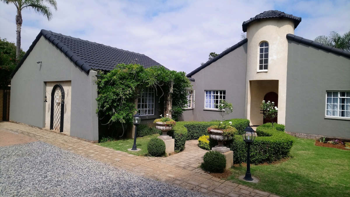 The exterior of the flat showcases a charming facade with a unique turret, surrounded by carefully landscaped gardens. Pathways made of stone lead to the entrance, flanked by manicured bushes and decorative planters. A mixture of green lawns and gravel adds texture to the outdoor space.