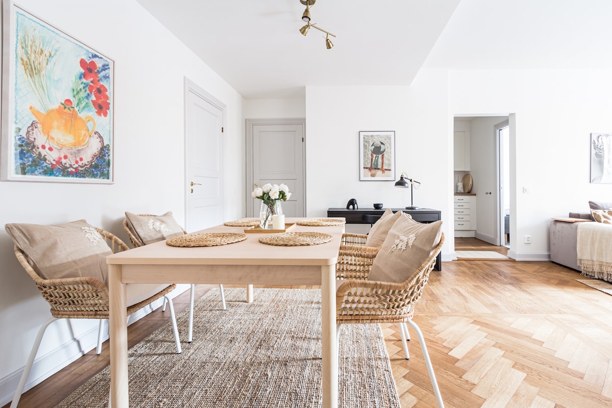 An open-plan dining area features a light wood table surrounded by four rattan chairs. A neutral rug covers the hardwood floor. A framed art piece adorns the wall, and natural light brightens the space, enhancing the soft tones of the decor.