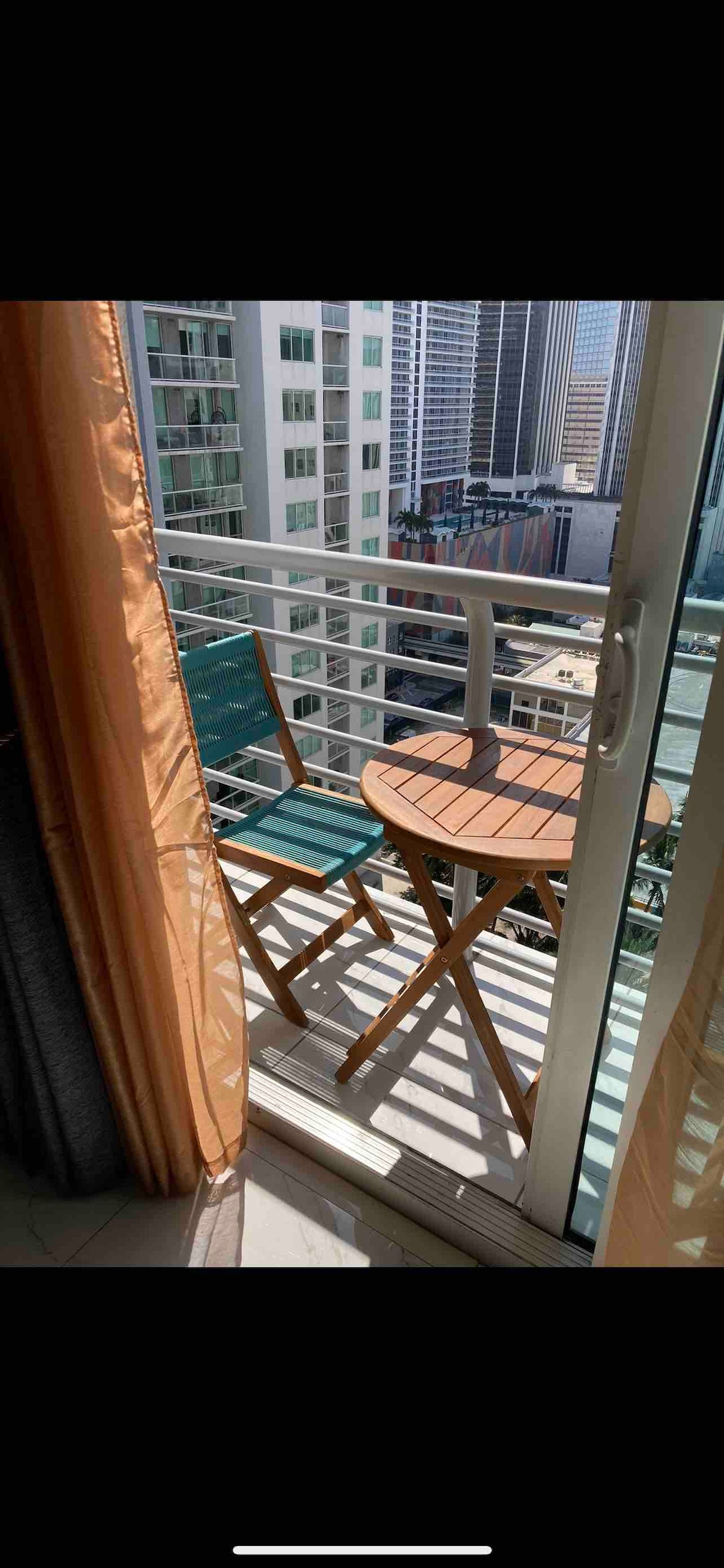A small balcony is shown with a round wooden table and two folding chairs. Floor-to-ceiling glass doors open to the outdoor space, providing a view of the surrounding city skyline and buildings, allowing natural light to enter the interior.