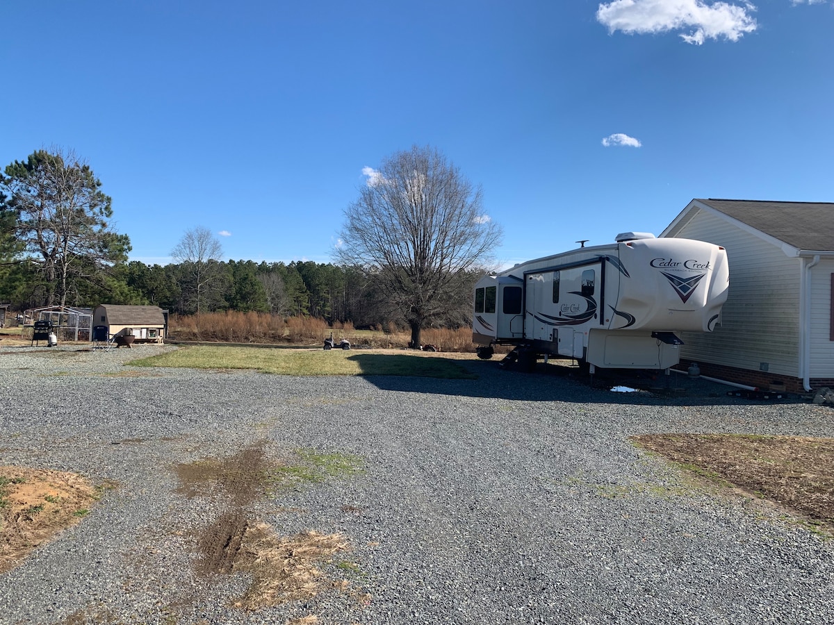 A spacious gravel area beside a home accommodates an RV, with a clear view of open fields and trees in the background. The sky is bright and blue, with a few scattered clouds, creating a peaceful rural setting.