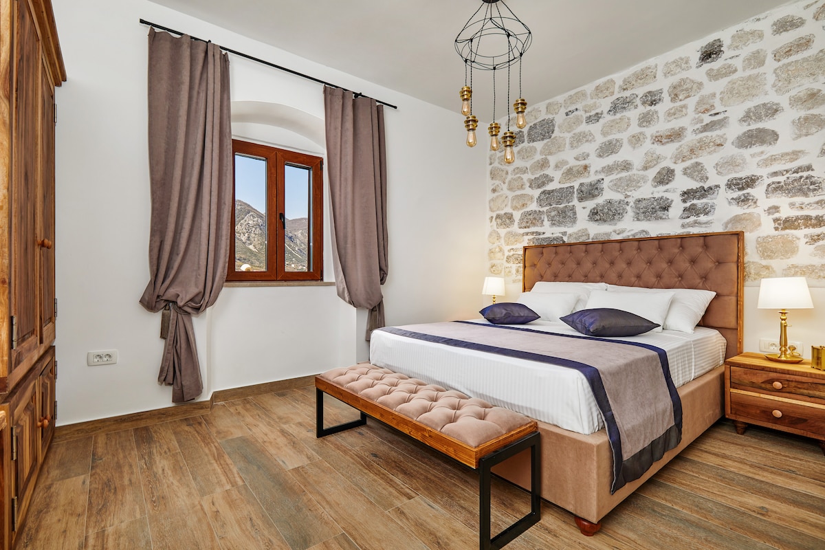 A spacious bedroom features a large bed with plush bedding, complemented by contrasting throw pillows. A wooden bench sits at the foot of the bed. Soft drapes frame a window that offers a view of the surrounding mountains, while warm lighting is provided by bedside lamps.