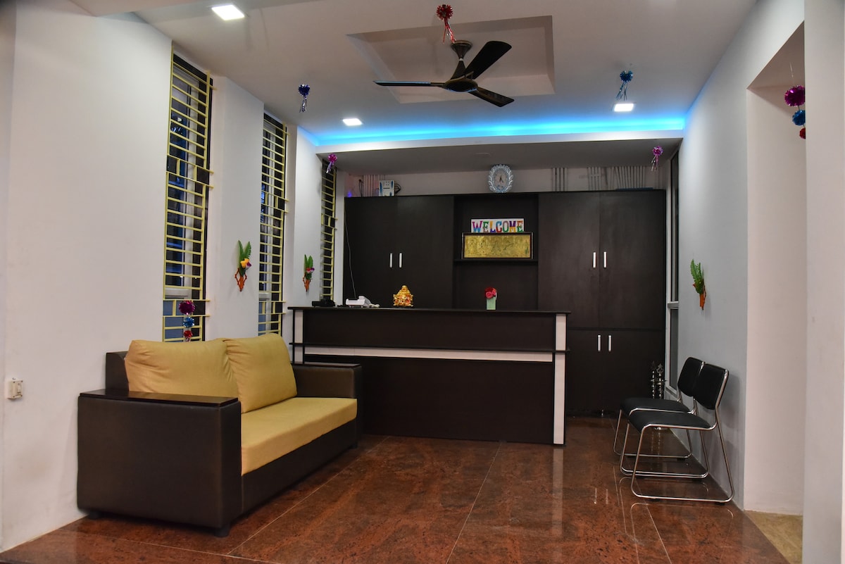 A reception area is presented with a dark wooden counter and a comfortable two-tone sofa. The space features walls adorned with decorative elements, while a ceiling fan and soft lighting enhance the ambiance. Large windows allow natural light to fill the area.