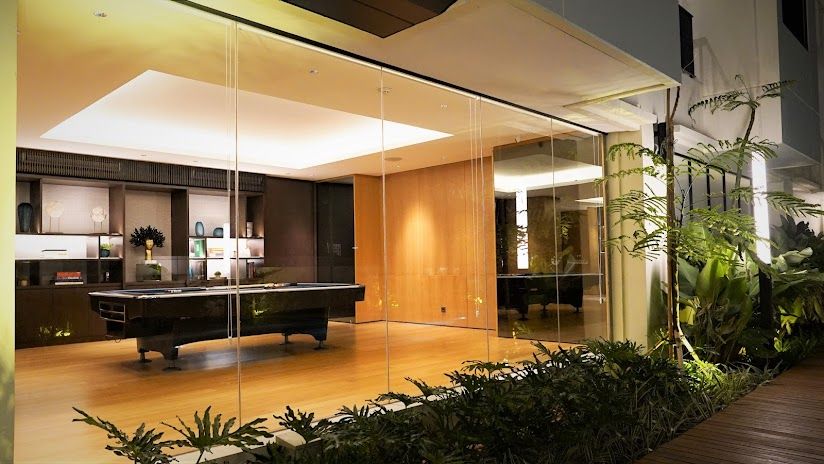 A spacious lounge area is enclosed by large glass panels, allowing natural light to illuminate the wooden flooring. Modern furnishings are arranged thoughtfully, complementing the sleek design. Lush greenery surrounds the exterior, enhancing the tranquil atmosphere.