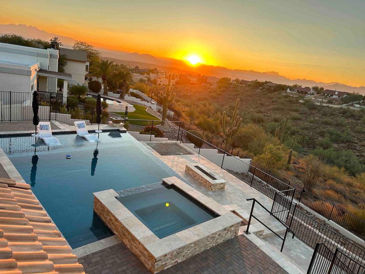 The image captures a serene outdoor setting at sunset, featuring a pool with a spa. The pool area is surrounded by a fence, and lounge chairs are positioned nearby. The landscape showcases distant mountains and cacti with the warm colors of the sky reflecting on the water.