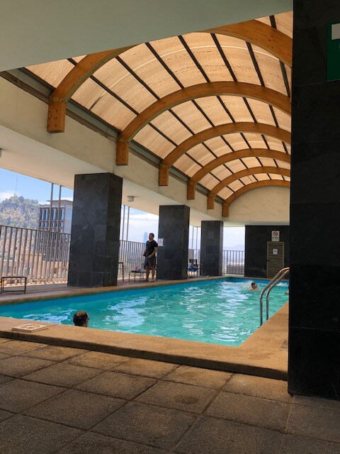 An indoor swimming pool is shown, featuring a clear blue water surface. Large windows allow natural light to fill the space, highlighting the wooden beam ceiling. A few individuals are seen enjoying the pool, while poolside lounging areas offer room for relaxation.