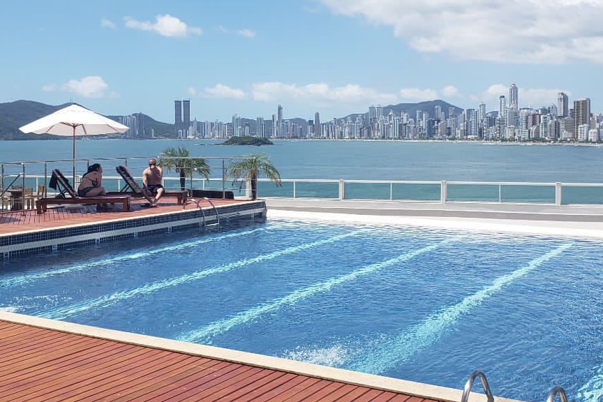 A pool area is displayed with a blue swimming pool featuring clear water. Lounge chairs are positioned by the edge, accompanied by a shaded umbrella. In the background, a city skyline overlooks the water, creating a scenic view under a partly cloudy sky.