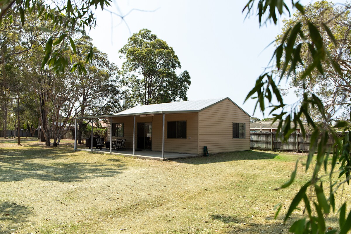 A spacious two-bedroom apartment is set within a rural landscape, showcasing a covered verandah that leads to a green lawn. Surrounding trees provide shade, while large windows invite natural light into the living areas. The exterior features a neutral color palette, enhancing the serene environment.
