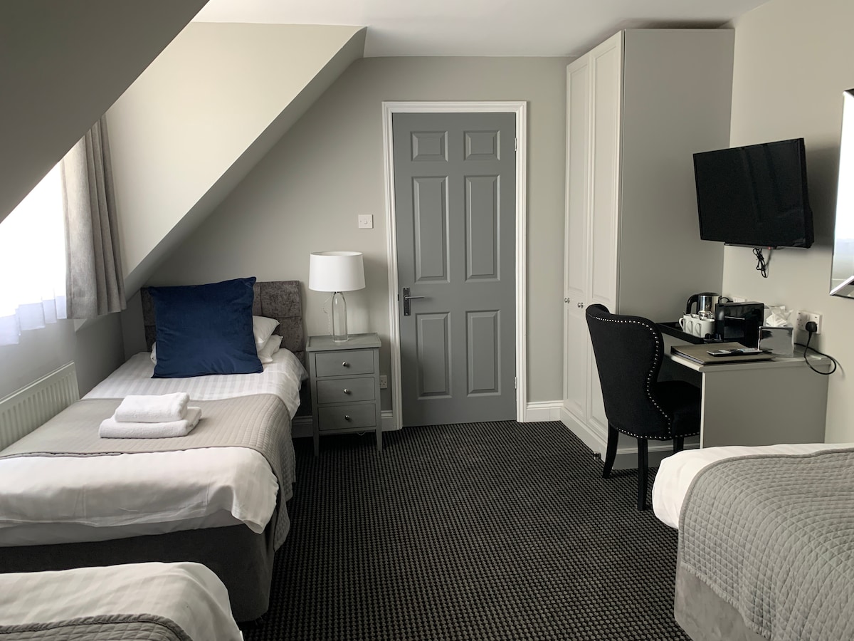 A well-appointed triple room features three single beds with soft bedding. A small side table is positioned beside one bed, and a dark chair is placed at a writing desk. A flat-screen TV is mounted on the wall opposite, with natural light filtering through the curtains.