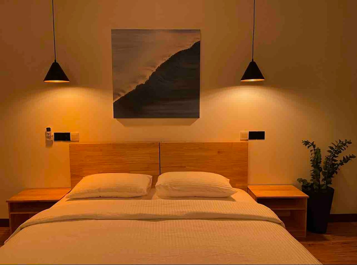 A cozy sleeping area features a spacious bed with light linens and two wooden nightstands. Soft lighting is provided by pendant lamps on either side. A piece of artwork depicting a wave is displayed above the headboard, complemented by a potted plant near the wall.