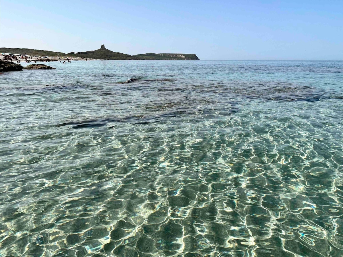 Clear turquoise water is visible, reflecting the sunlight and creating patterns on the surface. The shoreline is lined with soft sand and rocky outcrops, while a distant land formation can be seen against the blue sky, enhancing the serene coastal landscape.