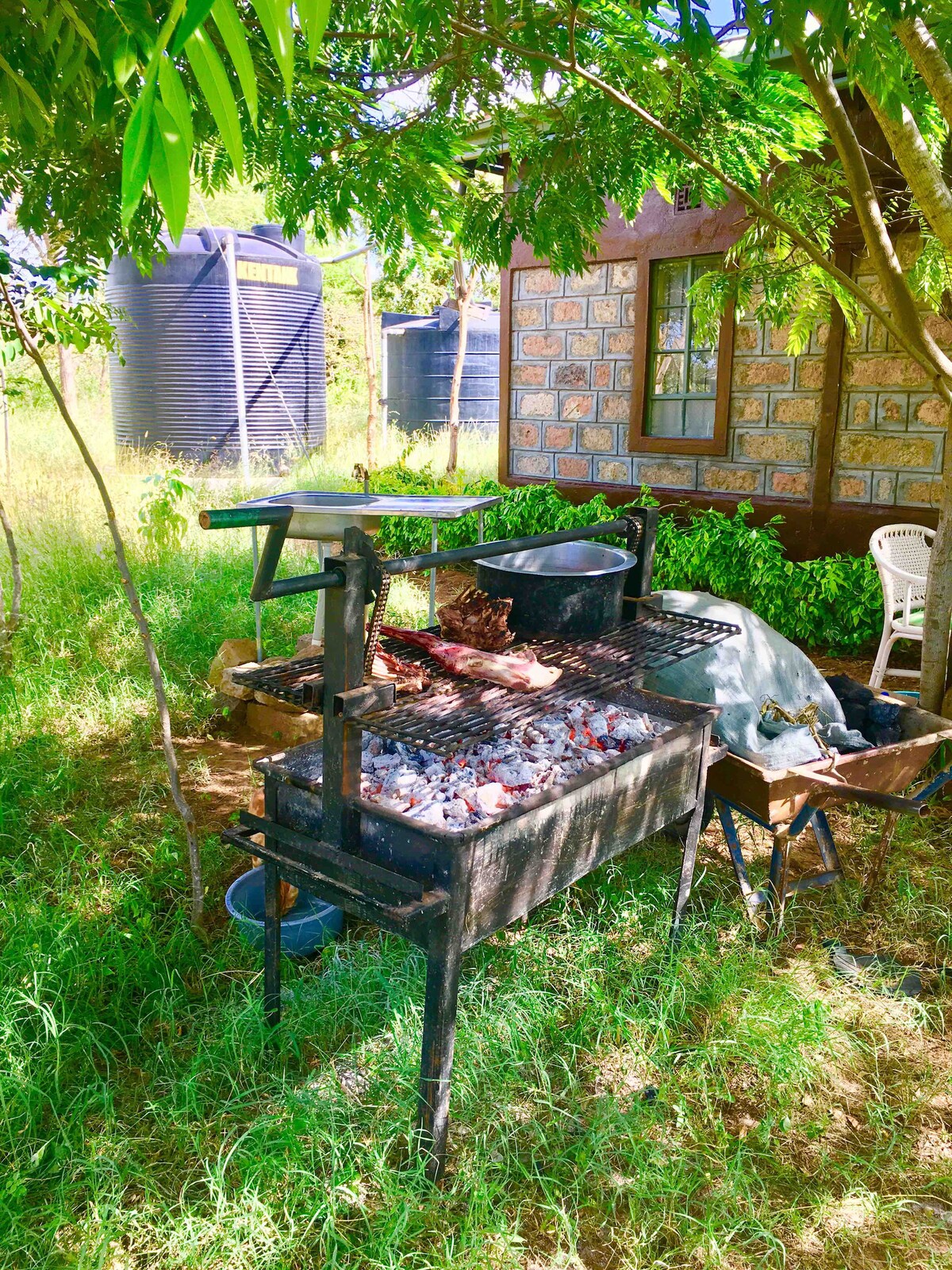 A charcoal grill is situated beneath a leafy tree, surrounded by lush grass. Water tanks are visible in the background, and a white chair is stationed nearby. The grill features a cooking surface with charcoal, ideal for outdoor meal preparation.
