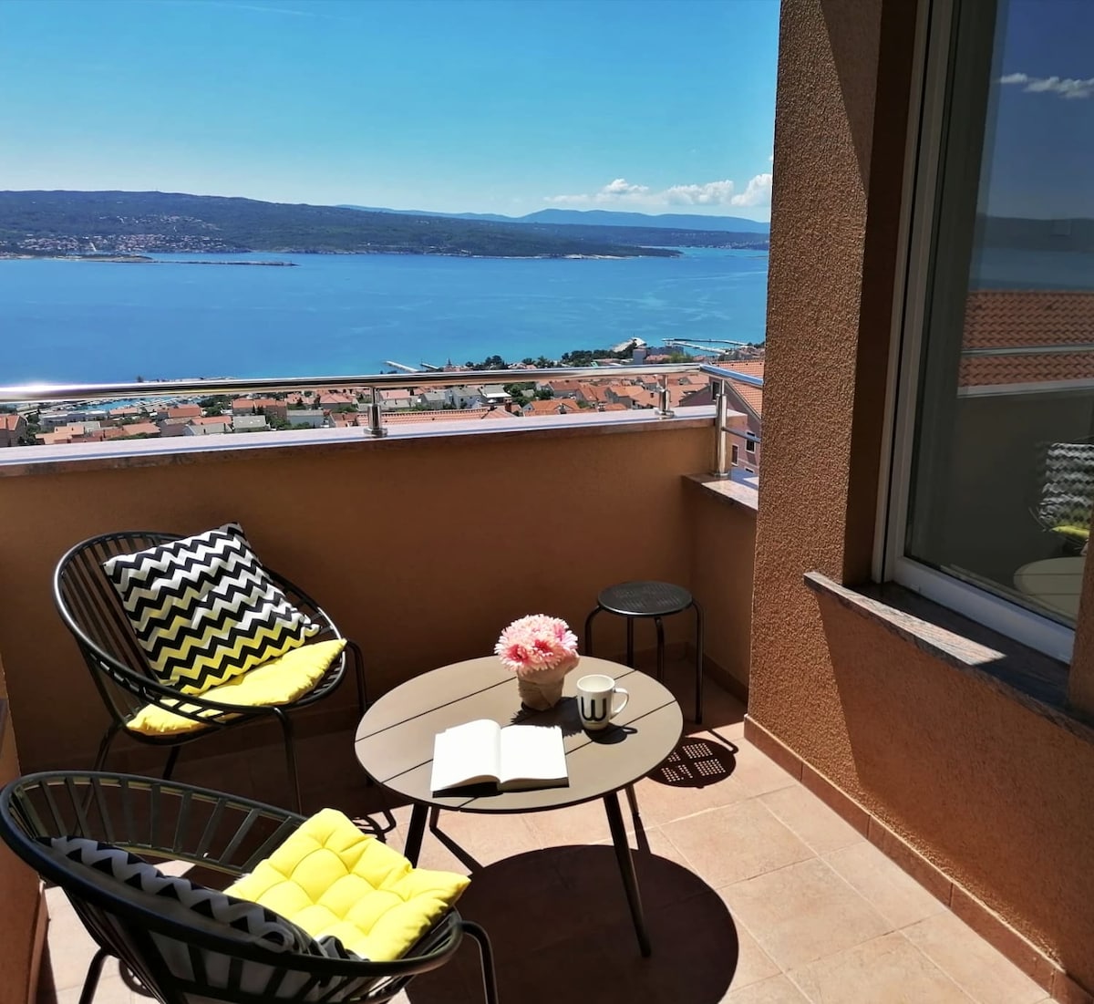 A balcony setting features two black chairs with bright yellow cushions and a small table. A flower arrangement adds a touch of color. The expansive view of the sea and surrounding landscape is visible beyond the balcony railing, with a clear blue sky above.