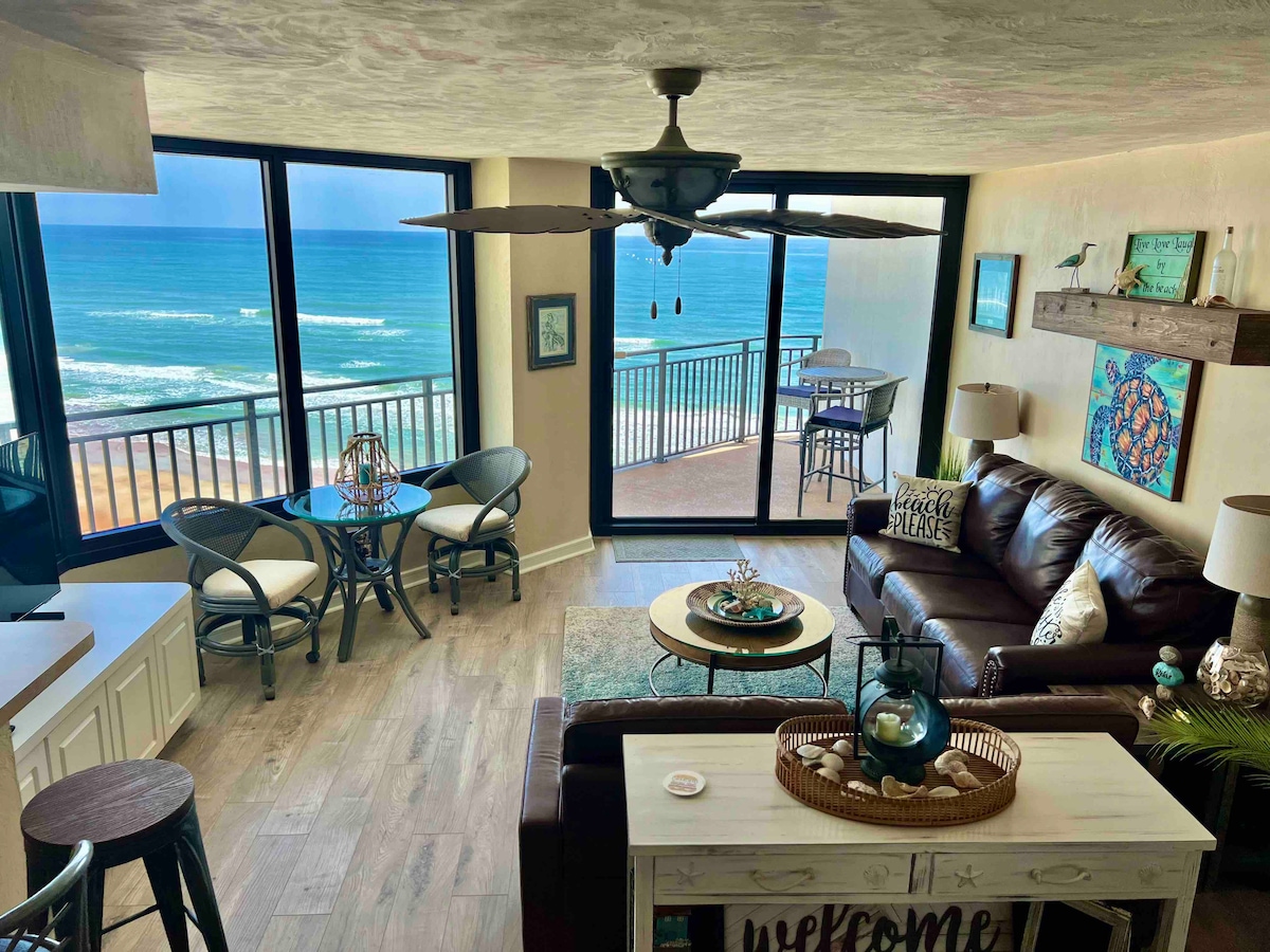 Spacious living area features large windows providing sweeping ocean views. A dark leather sofa and chairs surround a coastal-themed coffee table, while a dining table for four is situated near the balcony doors. A ceiling fan adds comfort to the inviting space.