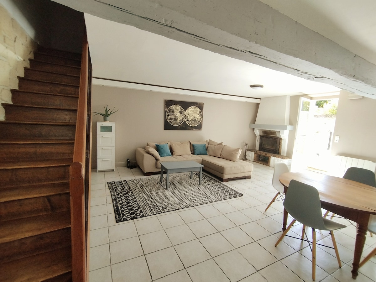 A spacious living area features a light-colored sectional sofa, a small coffee table, and a dining table surrounded by chairs. Natural light floods the room through large windows, highlighting a patterned rug on the tile floor. A staircase leads to the upper level.