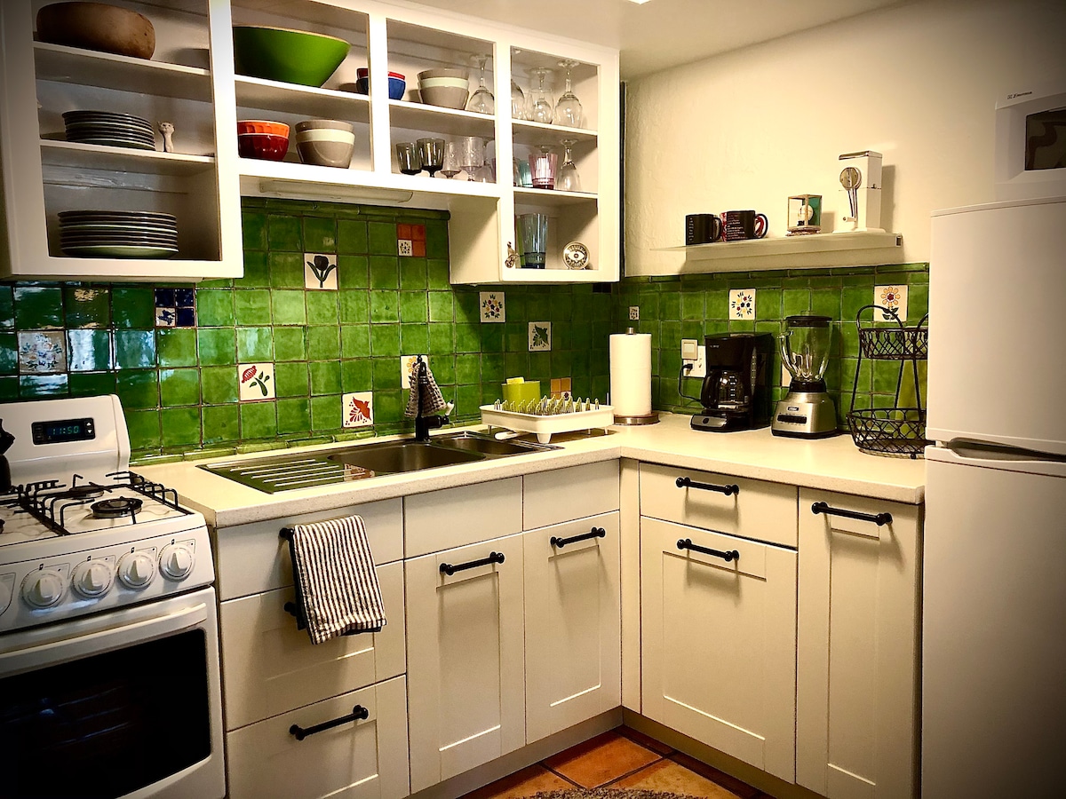 A bright kitchen is featured with green tile accents and open shelving displaying various dishes and glassware. Essential appliances include a gas stove, refrigerator, and coffee maker. Counter space is available for meal preparation, while a dish rack and blender are also present.
