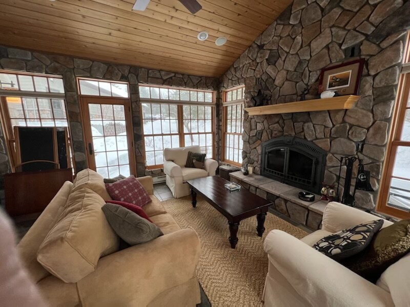 A stone-walled enclosed porch features large windows allowing natural light to fill the space. Comfortable seating is provided by soft sofas, centered around a wooden coffee table. A fireplace is positioned against the far wall, enhancing the cozy atmosphere of the room.