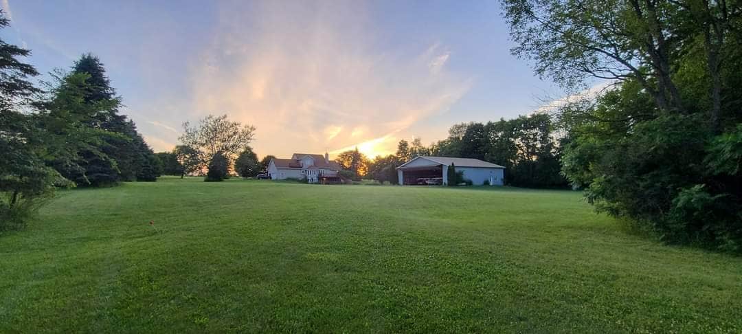 A spacious green yard stretches toward a sunset, with the house and garage visible in the background. Trees frame the scene, adding depth to the landscape. Soft clouds illuminate the sky in warm hues, creating a tranquil ambiance over the property.