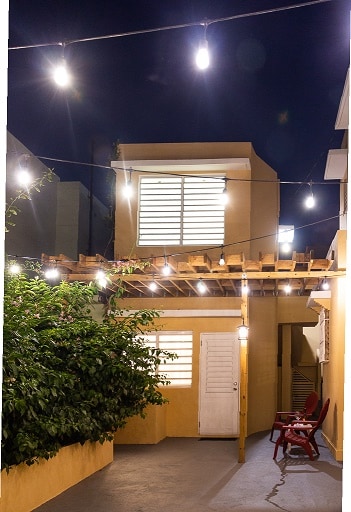 A charming outdoor space is illuminated by string lights, creating a warm ambiance at night. The yellow walls of the adjoining structure contrast with the greenery nearby. Two red chairs are positioned on the porch, inviting relaxation in this cozy setting.