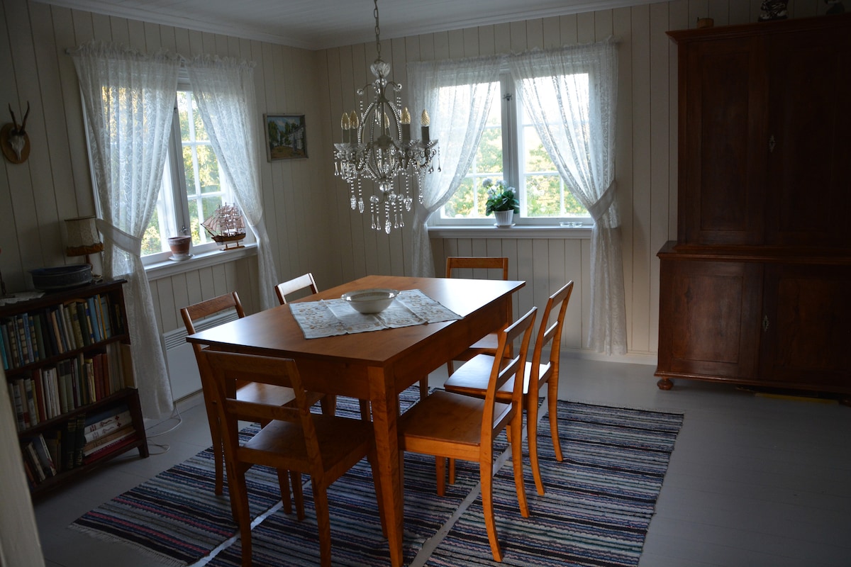 A dining area features a wooden table surrounded by four chairs, illuminated by natural light from two windows adorned with lace curtains. A decorative chandelier hangs from the ceiling, and a bookshelf is visible in the corner, enhancing the room's inviting feel.