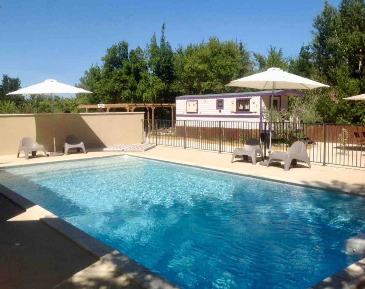 A refreshing swimming pool is surrounded by a secure fence, offering ample space for relaxation. Sun loungers are positioned beside the water, and large umbrellas provide shade. Lush greenery is visible in the background, with a roulotte situated nearby, enhancing the relaxing outdoor setting.