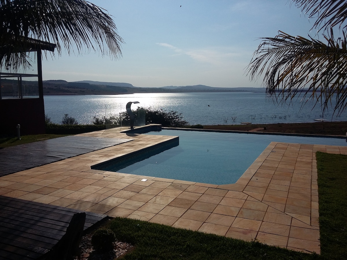 An inviting infinity pool reflects the sunlight, overlooking a serene body of water. The area is surrounded by lush greenery and features a wooden deck. Gentle ripples are visible on the water, enhancing the peaceful ambiance of the outdoor space.