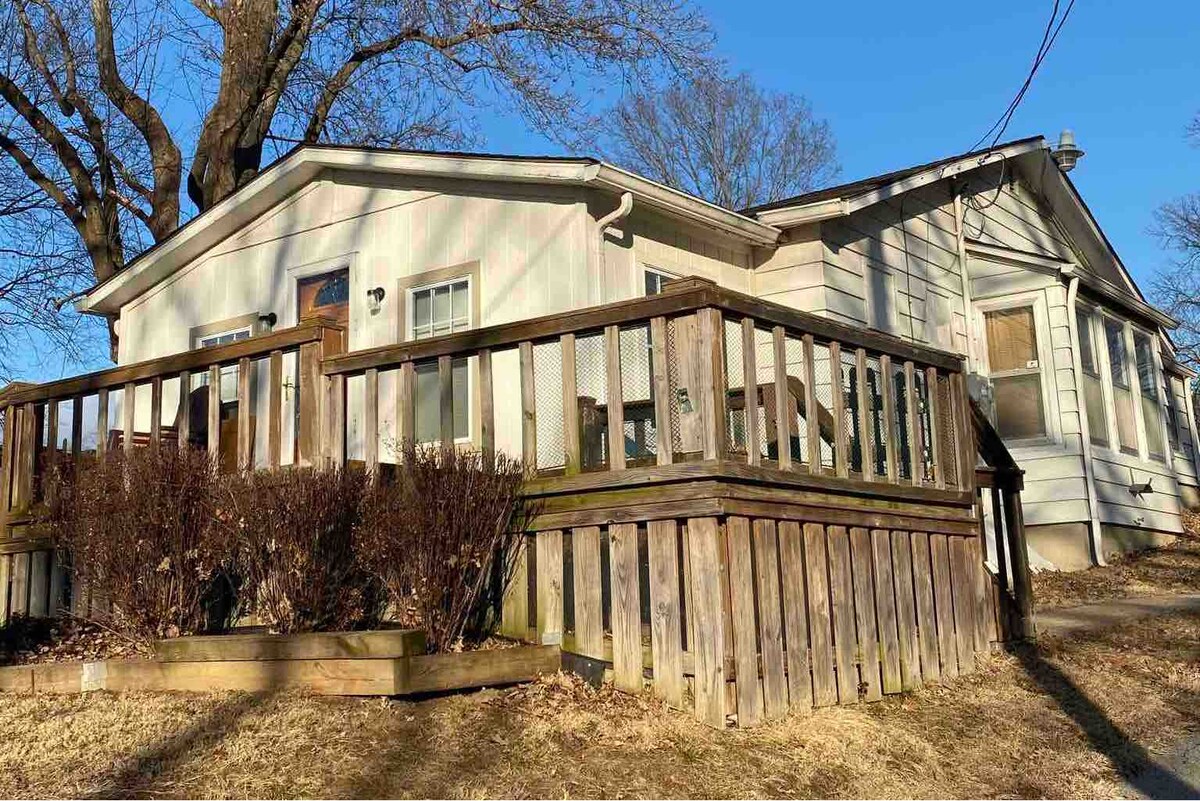 The exterior of a 1917 bungalow is displayed, featuring a wide wooden deck with a low railing. A compact garden area is positioned nearby, and the house is surrounded by mature trees. Sunlight brightens the scene, highlighting the home's charming architectural details.