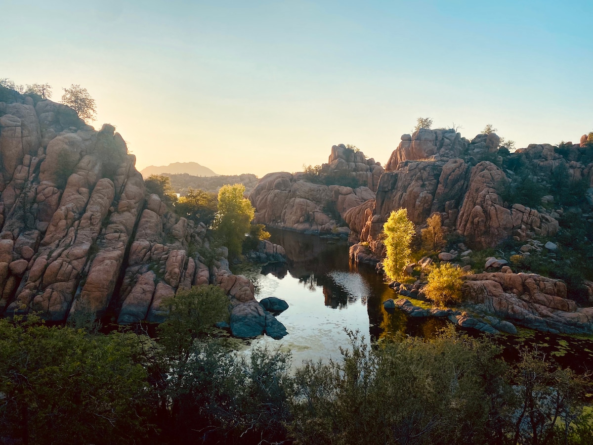 The image features a serene landscape of large, rugged rock formations surrounding a calm body of water. Lush greenery is present along the shores, with trees reflecting in the water at sunrise. Soft light highlights the natural beauty of the area, creating a peaceful atmosphere.
