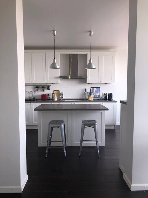 The kitchen as seen from the living room. The kitchen features a cooking zone, bar chairs, a double door fridge, a separate wine fridge, a microwave, an oven, induction cooker, a dishwasher, a Nespresso coffee machine, electric kettle, a toster, a juicer.