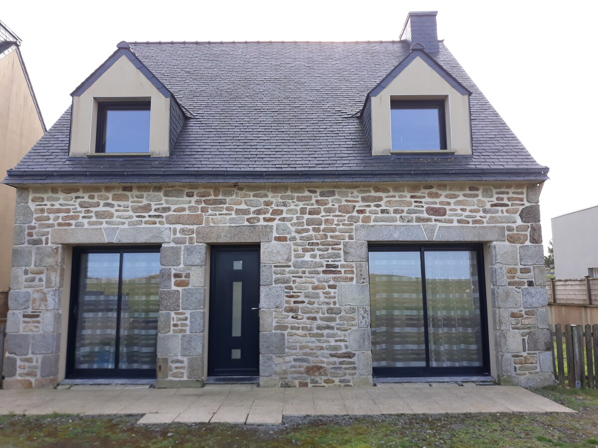 A charming stone house with a gabled roof features two large front windows framed by dark trim. The main entrance is centered and highlighted by a modern door. The exterior presents a blend of natural stone textures, complemented by a neat pathway in front.