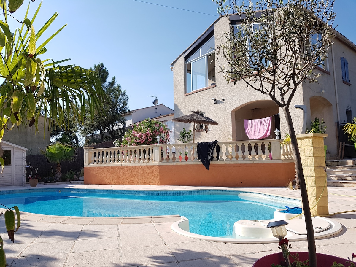 The outdoor area showcases a private swimming pool surrounded by a tiled patio. Lush plants, including a palm tree, enhance the serene setting. The villa's façade features large windows and a covered terrace. Laundry hangs on a line, adding a homely touch to the scene.