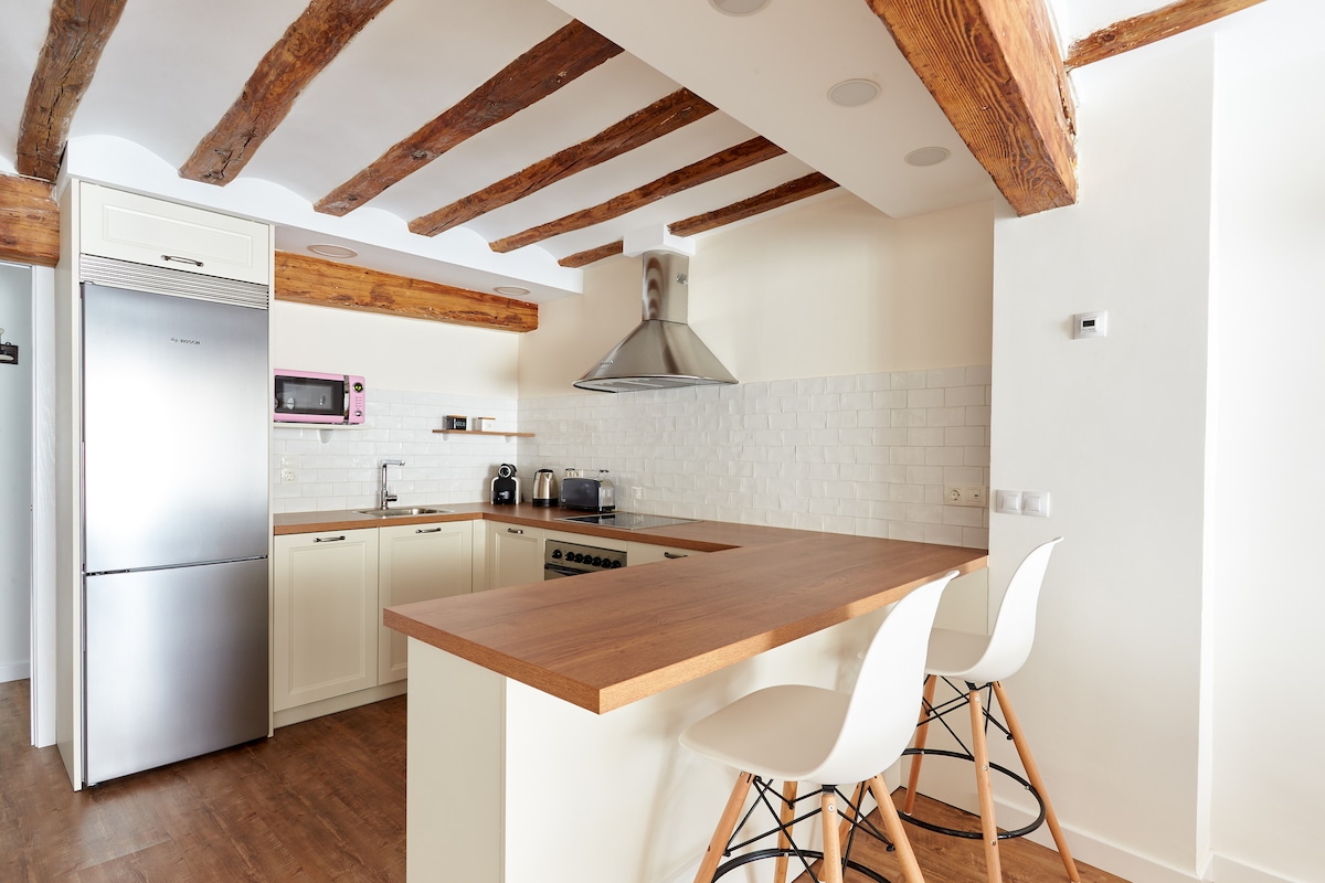 A modern kitchen is highlighted by wooden beams overhead and a spacious island with high-backed stools. Appliances include a refrigerator, microwave, and a sleek range hood above the stove. Light cabinetry complements the white tiled backsplash, creating a bright cooking space.