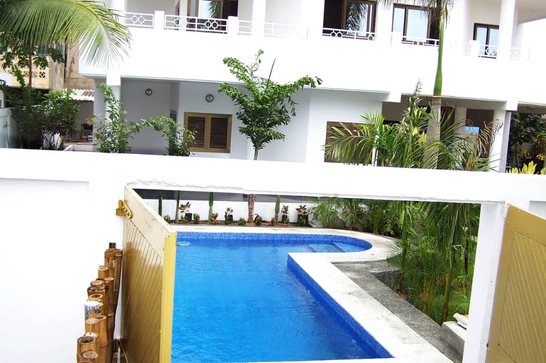A modern villa featuring a clear blue pool surrounded by lush greenery and well-maintained landscaping. The structure includes two levels with multiple windows allowing ample natural light. Decorative plants add a touch of nature to the serene outdoor area.