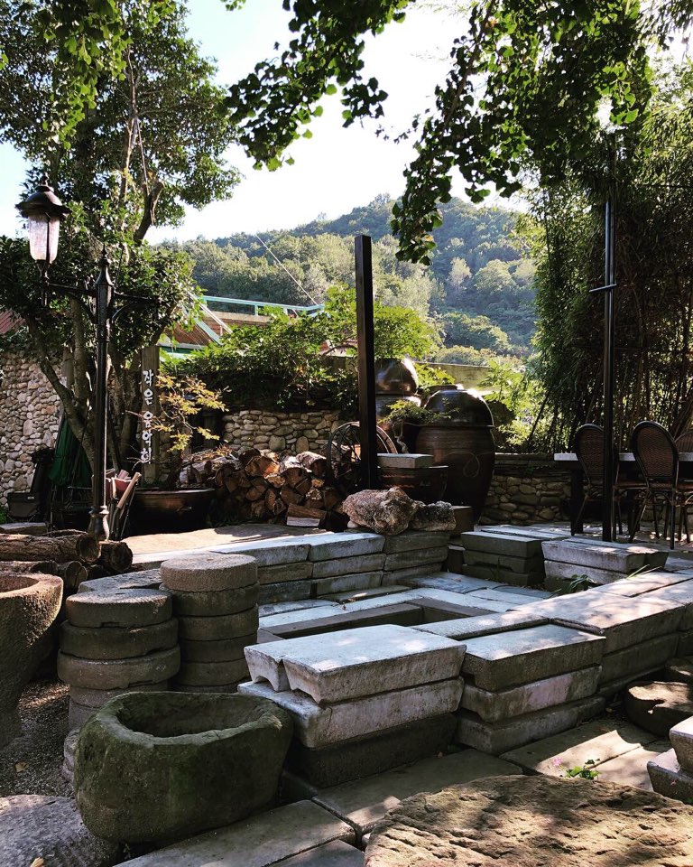 A serene outdoor space features stacked stone slabs and circular stone planters. Lush greenery surrounds the area, with trees providing dappled sunlight. In the background, wooden logs are neatly arranged, and a set of dark metal chairs and a table are seen nearby.