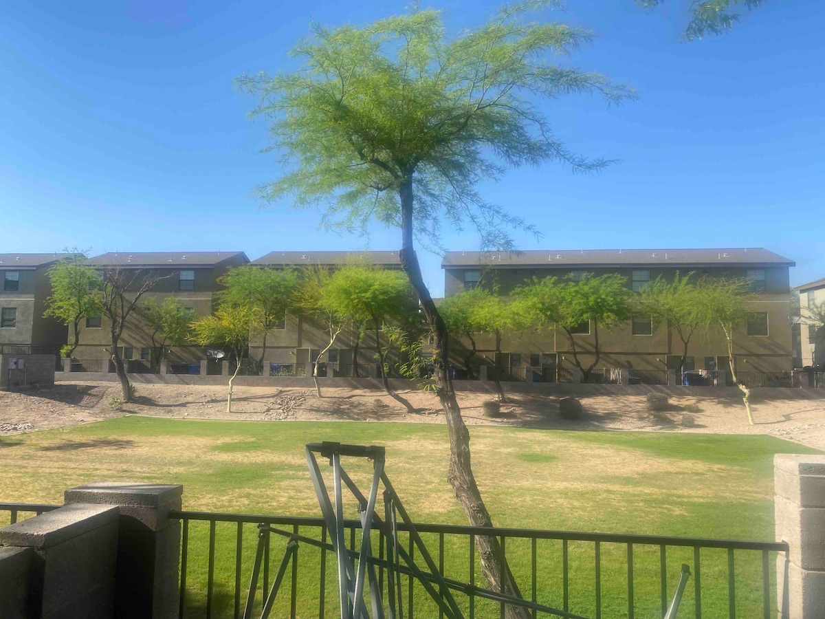 A view of a grassy area framed by a green fence, showcasing several trees and low shrubs. Behind the green space, a series of two-story buildings are visible, each featuring multiple windows. The sky is clear and blue, contributing to a bright outdoor setting.