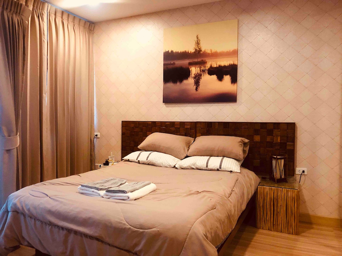 A spacious bed with soft bedding is positioned against a textured wall. Light curtains frame the window, allowing soft illumination. A piece of wall art depicting a tranquil landscape is centered above the bed, while a small side table adds utility to the serene space.