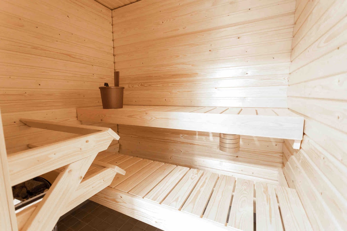 A wooden sauna interior is depicted, featuring light-colored wooden walls and benches. The space includes a wooden bucket and ladle on a shelf, with multiple wooden benches arranged for relaxation. Natural light highlights the clean lines and warm tones of the wood.