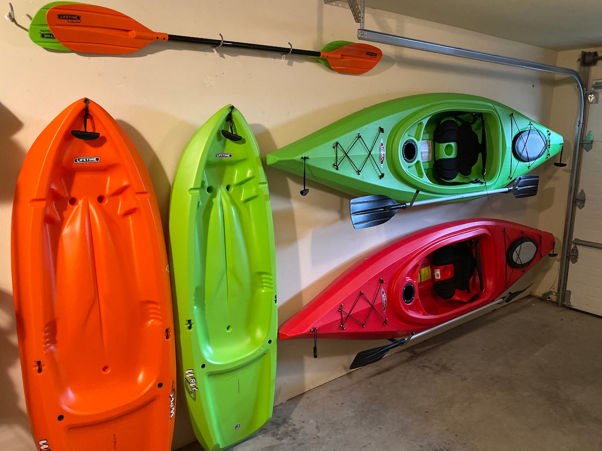 Two brightly colored kayaks rest against the wall, one orange and one red, alongside a green kayak with black accents. A kayak paddle in vibrant orange and green is mounted above, emphasizing the variety of water sports available.