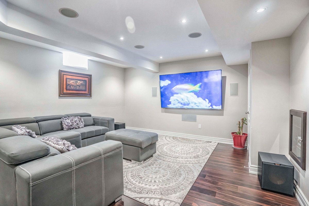 A spacious living area is equipped with a large sectional sofa, complemented by a soft area rug. A flat-screen television is mounted on the wall, while recessed lighting enhances the overall brightness of the room. A small plant adds a touch of greenery.
